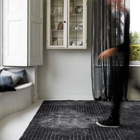 Eine verschwommene Gestalt läuft über einen dunkel gemusterten Teppich in einem minimalistischen Raum mit grauen Wänden