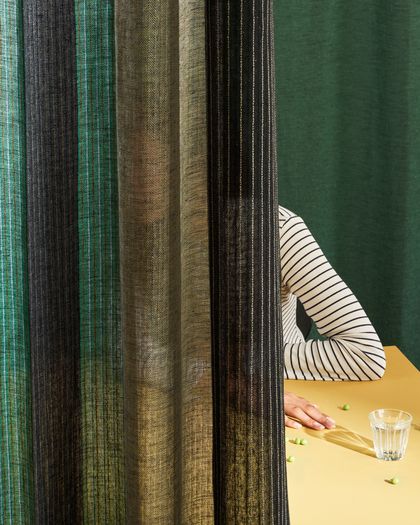 Eine Person in einem gestreiften Hemd sitzt an einem gelben Tisch mit einem Glas Wasser, die teilweise von durchsichtigen Vorhängen verdeckt werden.